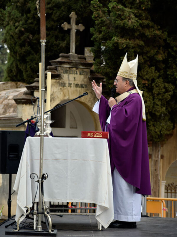 Un sacerdote en hábito púrpura ofrece un sermón en un entorno al aire libre, con un altar y una cruz en el fondo.