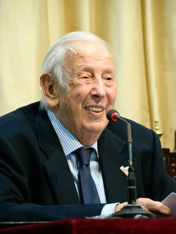 Senior man in formal attire speaking at a podium with a microphone.