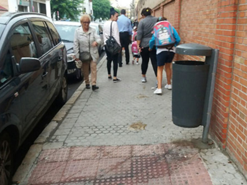 Gente caminando por una calle urbana junto a un coche estacionado y un contenedor de basura junto a un muro de ladrillo.