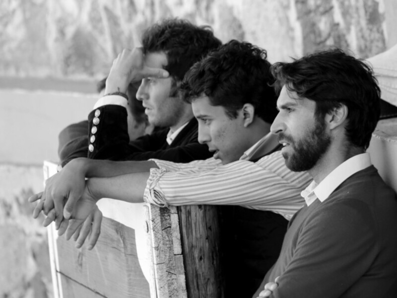 Tres hombres sentados juntos, apoyados en una estructura de madera, en un entorno al aire libre con paredes de piedra en el fondo.