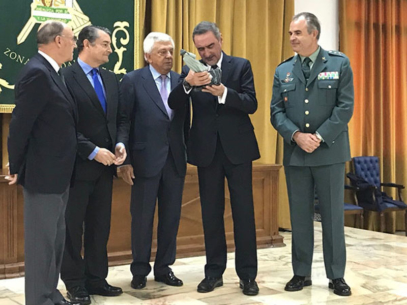 Cuatro hombres en trajes formales, uno de ellos en uniforme militar, posando para una foto en un entorno formal con cortinas y un escudo en el fondo.