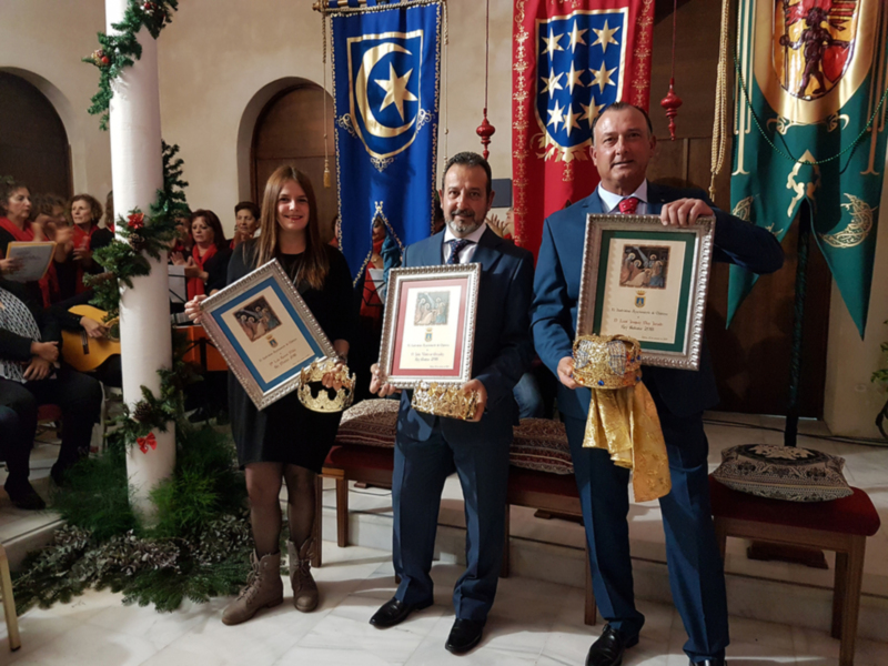 Tres personas posan con premios en una ceremonia formal, rodeadas de banderas y decoración navideña.