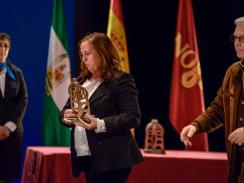 Una mujer recibe un premio mientras un hombre la felicita. Detrás de ellos, banderas de España y Andalucía ondean en el fondo.