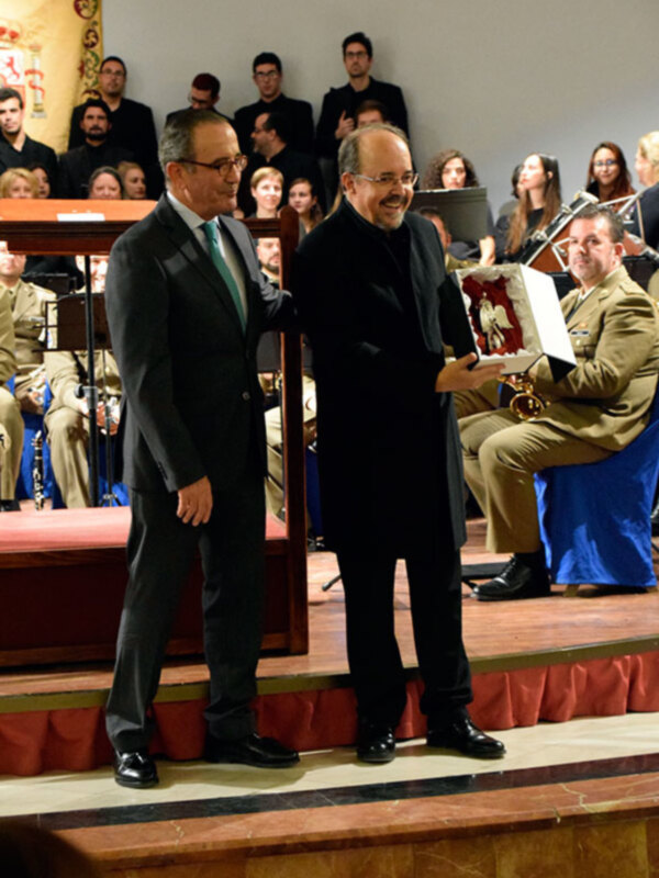 Dos hombres en trajes se encuentran en un escenario, uno de ellos sostiene un regalo. La audiencia se puede ver en el fondo, con algunos individuos vestidos con uniformes militares. El ambiente sugiere una ceremonia formal o un evento de reconocimiento.
