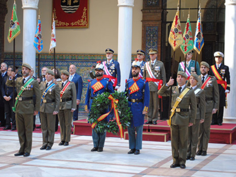 Desfile militar con oficiales en uniformes ceremoniales y guirnaldas de flores, ante un escudo y banderas decorativas.