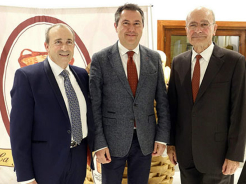 Tres hombres en trajes posan frente a una bandera con el logo de 'La Canasta' en un evento formal. El hombre en el medio lleva una corbata roja y los otros dos llevan corbatas azules. La escena sugiere una reunión o presentación de negocios.