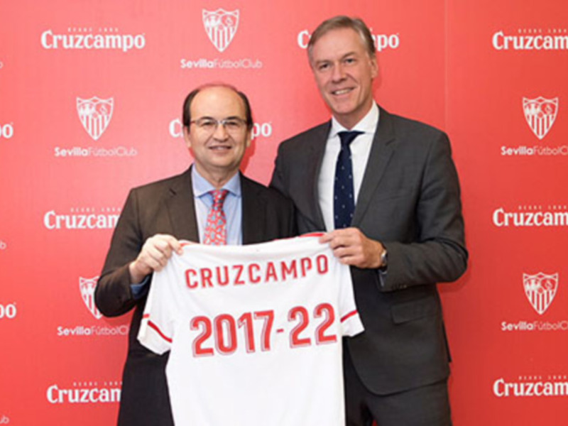 Dos hombres en trajes sostienen una camiseta del Cruzcampo con el año 2017-22 en un evento formal.