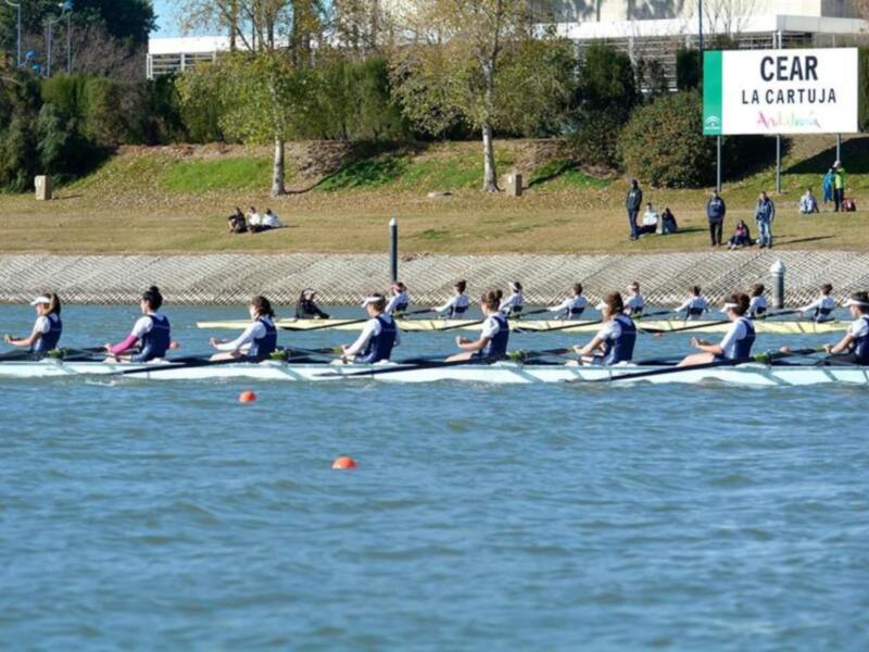 Equipo de remo en competencia en un río con espectadores en la orilla y un cartel de 'CEAR LA CARTUJA' en el fondo.