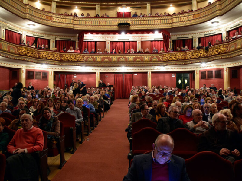 Interior de un teatro con audiencia sentada en butacas rojas, decoración ornamental y balcones con cortinas rojas.