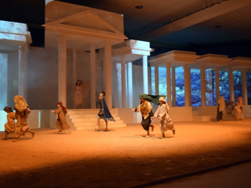 Recreación teatral de escena histórica con figuras en trajes antiguos y templo en el fondo.