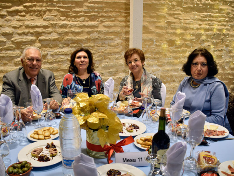 Personas disfrutando de una cena en una mesa decorada con flores doradas y copas de vino, en un ambiente acogedor con pared de ladrillo y luces de guirnalda.