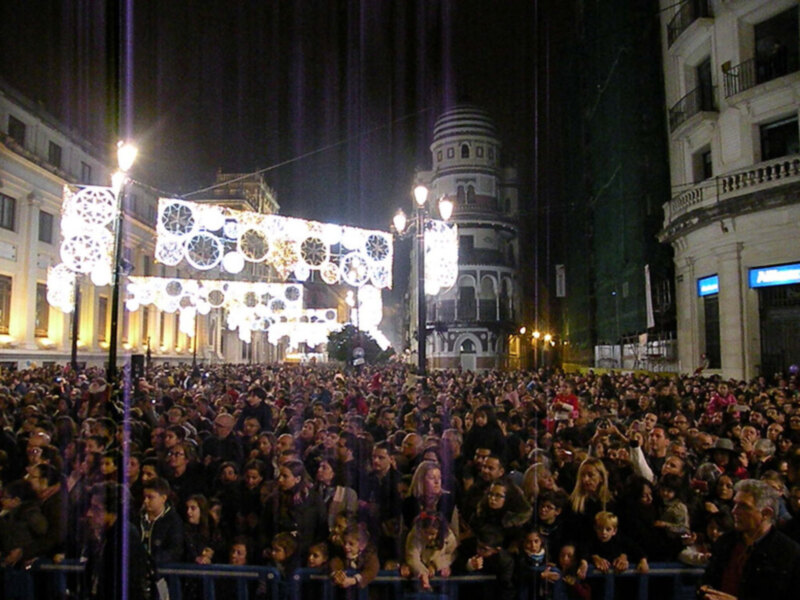 Una multitud de personas disfrutando de una celebración nocturna en una plaza con luces y decoraciones festivas. La arquitectura del edificio en el fondo destaca con su diseño único y la celebración parece ser una festividad importante en la ciudad.