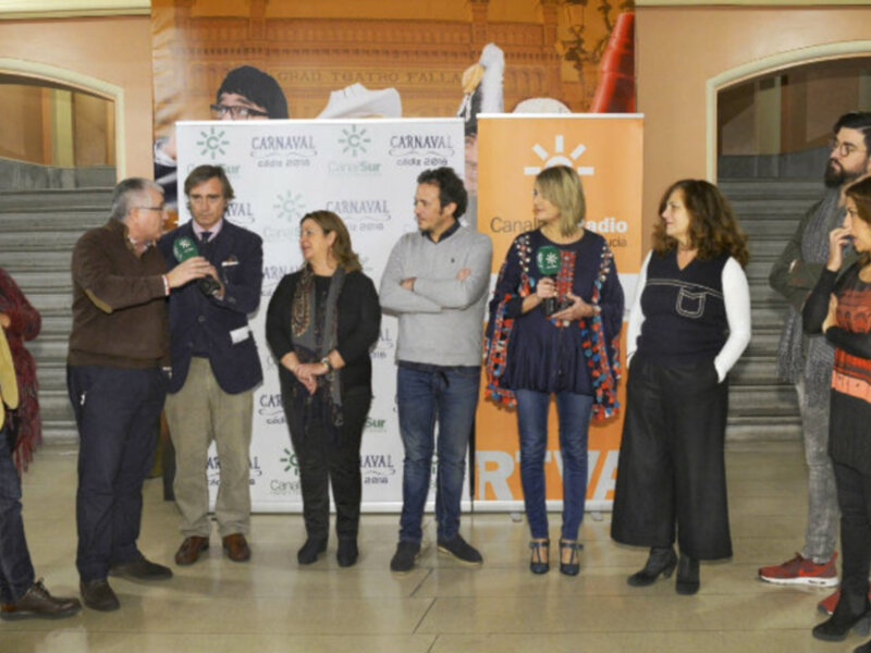 Presentación del Carnaval en un evento oficial con personas hablando y posando frente a carteles del Carnaval.