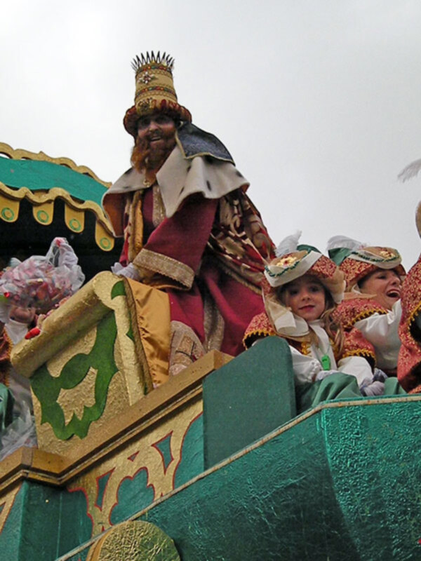 Un personaje vestido de rey medieval en un carro festivo, rodeado de personas con disfraces navideños.