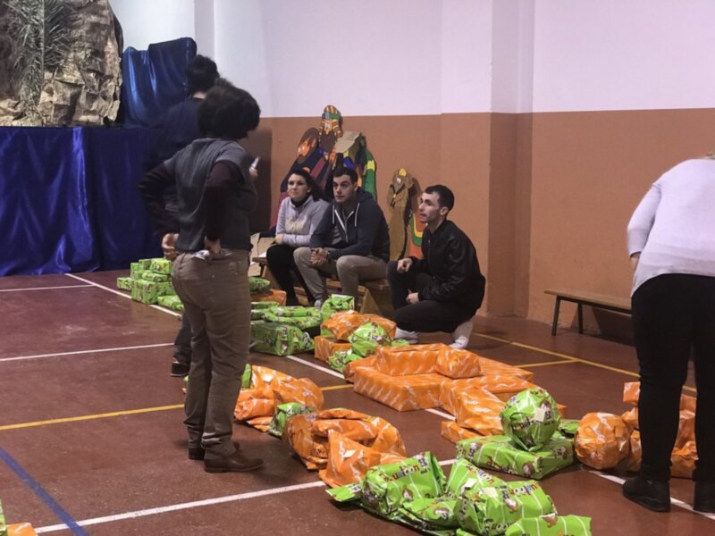 Un grupo de personas se reúne en un espacio interior, rodeado de paquetes de alimentos en bolsas de plástico. La pared del fondo está decorada con una figura de un personaje animado.