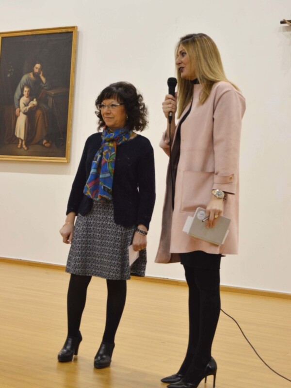 Dos mujeres en un evento, una con un abrigo rosa y la otra con un abrigo negro y una bufanda de colores brillantes, frente a un lienzo de arte en el fondo.
