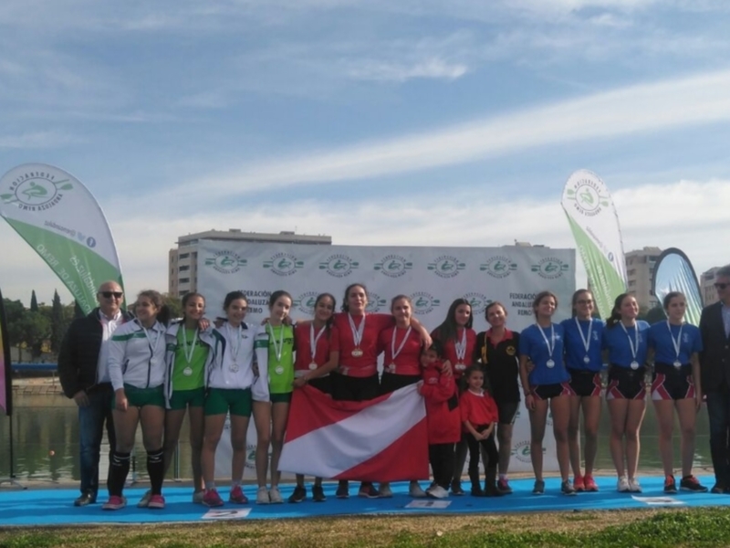 Equipo de remo femenino posando con medallas y bandera, celebrando un logro en un evento deportivo junto al río.