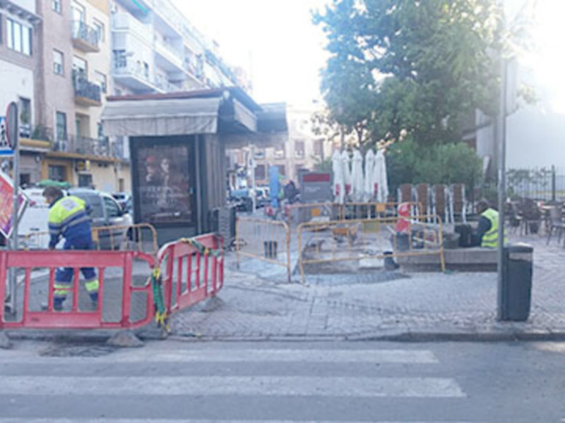 Trabajadores de mantenimiento en una calle con barreras de seguridad y un café al fondo.