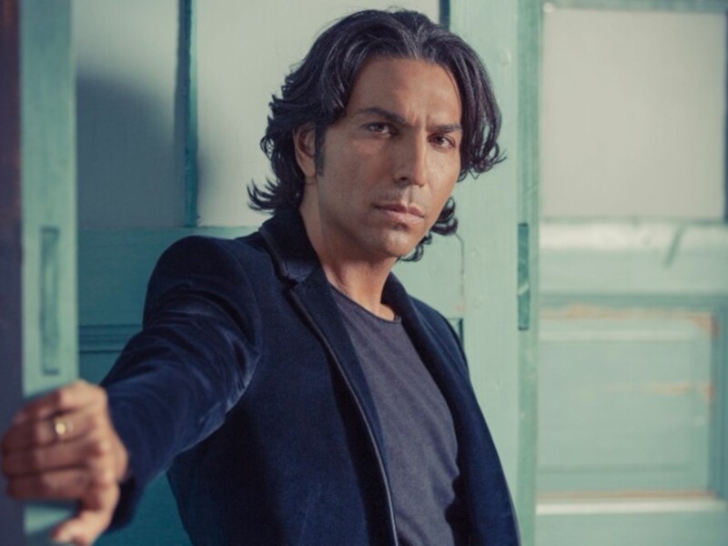 Un hombre con cabello largo y oscuro, vistiendo una chaqueta azul y una camiseta negra, apunta con su mano derecha hacia la cámara. El fondo es una puerta de madera verde con marcos blancos, lo que sugiere un ambiente interior.