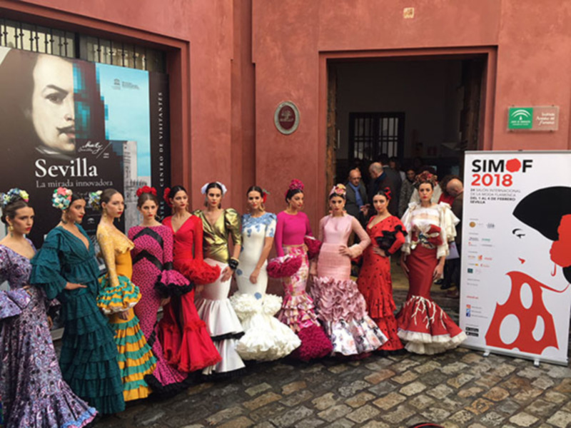 Modelos de trajes de flamenca posando frente a un edificio rojo, con un cartel de SIMOF 2018 en el fondo.