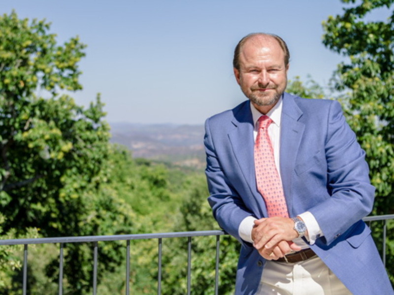 Hombre en traje azul y corbata, posando al aire libre con un paisaje verde y soleado en el fondo.