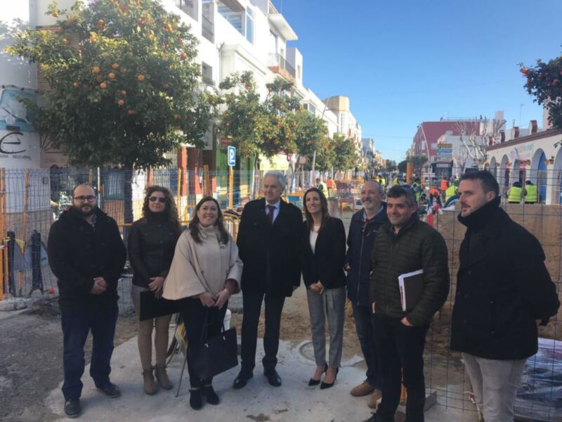 Oficiales y autoridades posan para una foto en un sitio de construcción en una calle con árboles frutales y edificios blancos.