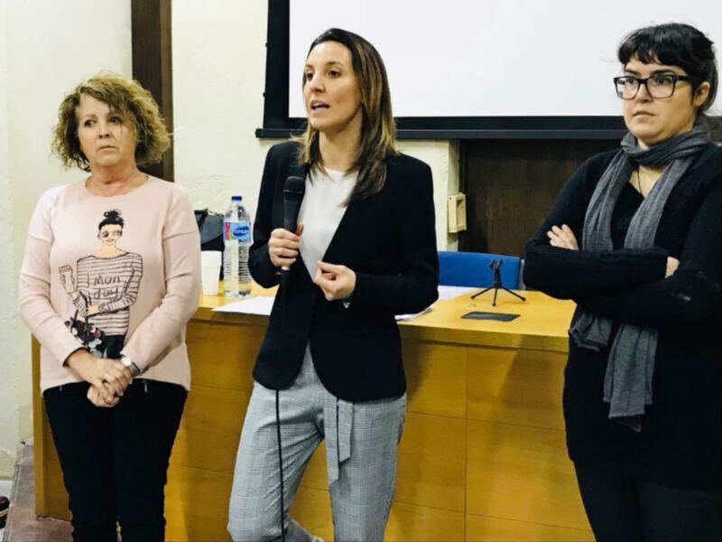 Tres mujeres participan en una reunión o presentación formal en un espacio con mesas y sillas.