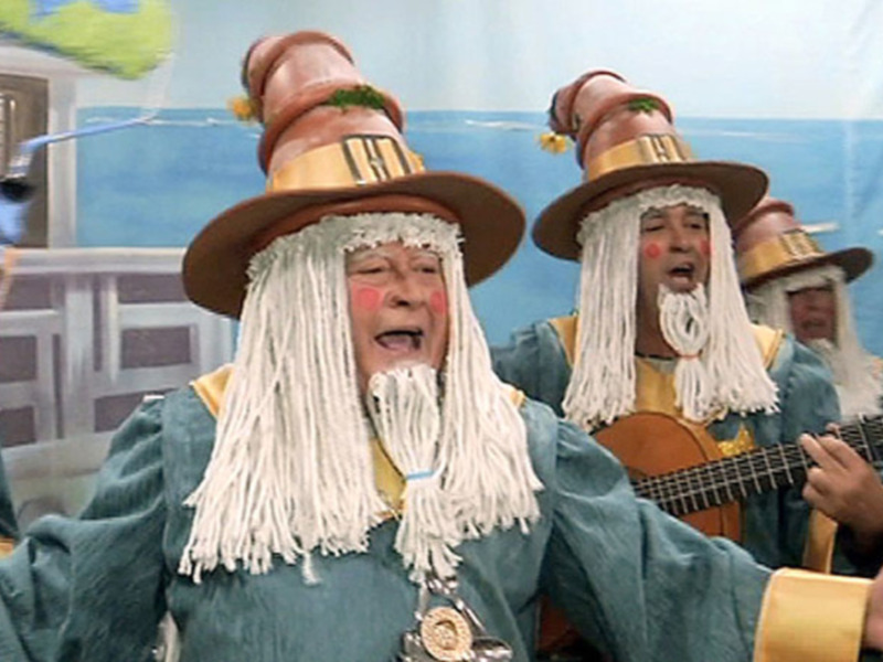Dos personajes con pelucas largas y sombreros coloridos, uno tocando una guitarra, en un entorno festivo con globos y decoraciones navideñas.