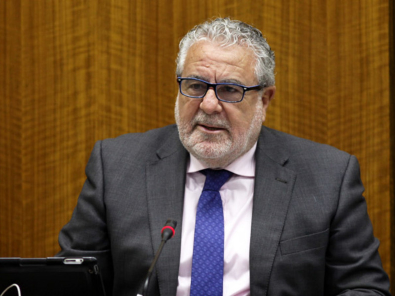 Un hombre de cabello blanco y barba, con gafas de montura oscura, hablando en un escenario formal. Se encuentra frente a una pantalla y detrás de él hay una mesa con botellas de agua. La pared del fondo es de madera de color claro.