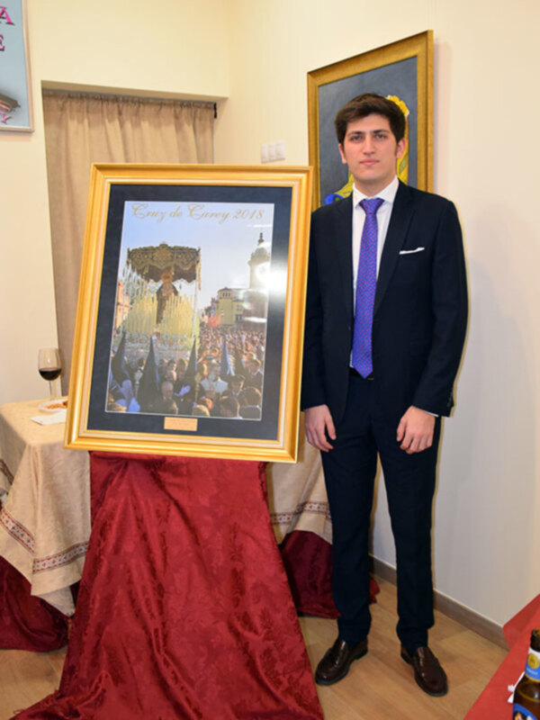 Un hombre en traje y corbata azul posa junto a una imagen de una procesión religiosa en un evento formal. La imagen muestra una multitud en una procesión con un edificio histórico en el fondo, y la fecha 2018 está escrita en la imagen.