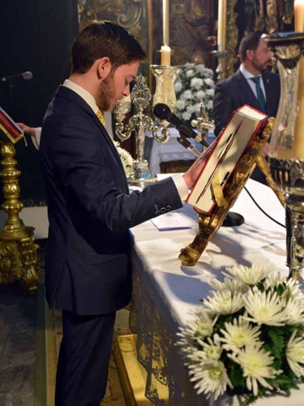 Persona leyendo de un libro en un entorno ceremonial con decoración barroca y flores blancas.