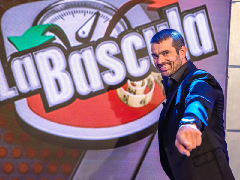 Personaje de 'Lo Básico' en un escenario con el logo del programa en el fondo.