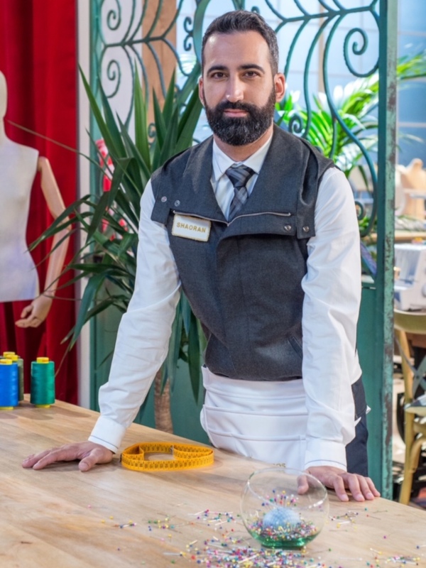 Un hombre vestido con un chaleco y corbata, posando detrás de una mesa de costura con pinzas y un metro.