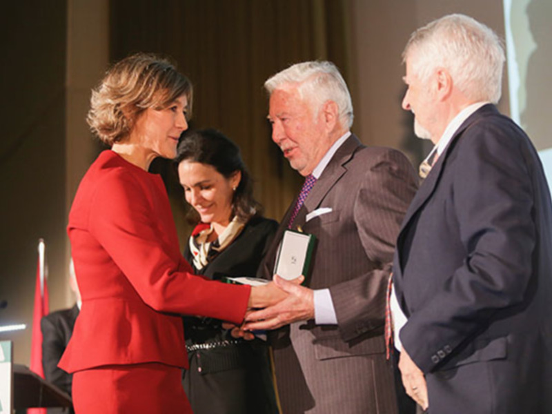 Entrega de premio en un evento formal, con una mujer en traje rojo entregando un reconocimiento a un hombre en traje gris.