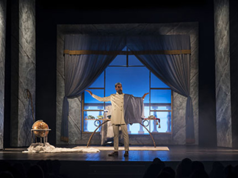 Actor on stage with arms outstretched, globe and props in the background, under a draped window.