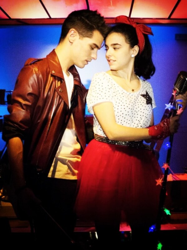 Dos personas vestidas con ropa retro, una con chaqueta de cuero roja y la otra con falda roja y top blanco, en un ambiente iluminado con luces de colores.