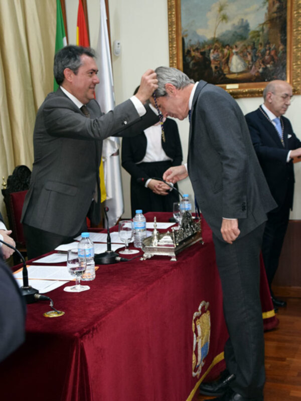 Un hombre recibe una medalla de manos de otro durante una ceremonia formal en un salón decorado con banderas y pinturas históricas.