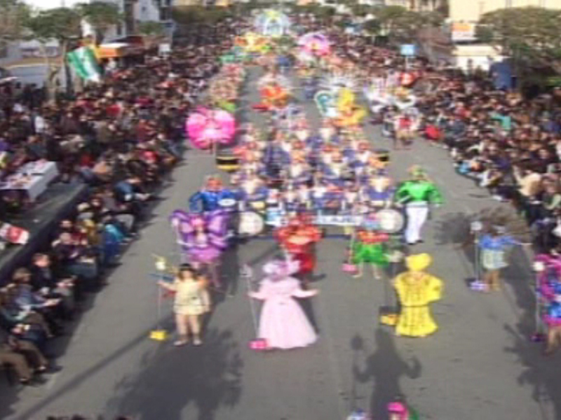 Desfile de disfraces en una calle abarrotada de público, con participantes vestidos de colores brillantes y figuras animadas.