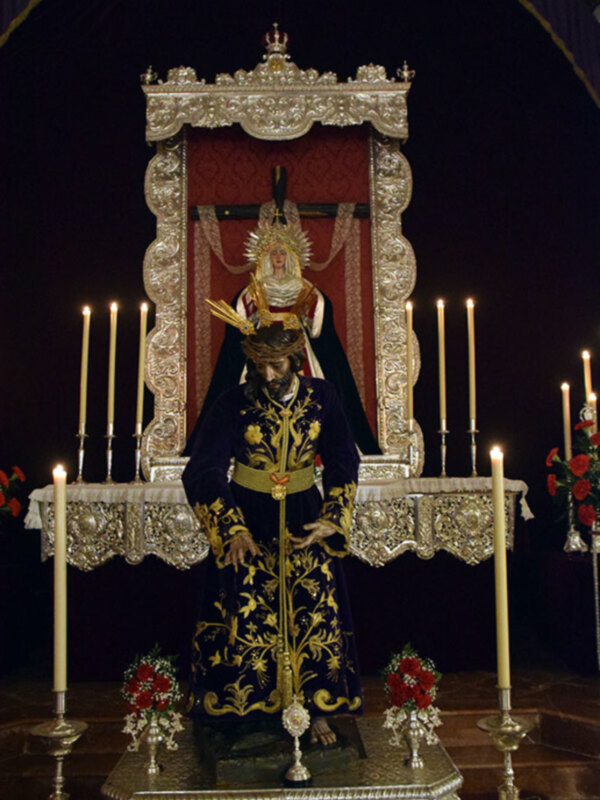 Estatua religiosa en un altar con velas y flores, iluminada por una araña de luz.