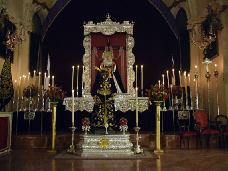 Altar con imagen procesional en un templo, adornado con candelabros y flores, bajo una estructura dorada y roja.