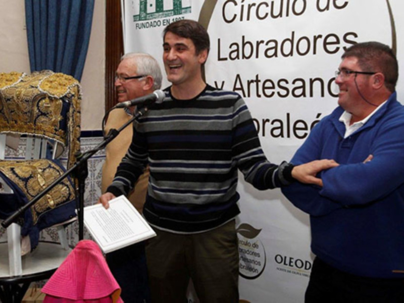 Dos hombres sonríen mientras uno de ellos sostiene un documento en una conferencia o evento público.