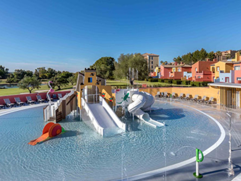 Piscina infantil con toboganes y juegos de agua en un entorno verde y soleado. El paisaje incluye edificios y árboles en el fondo, creando un ambiente familiar y relajante.