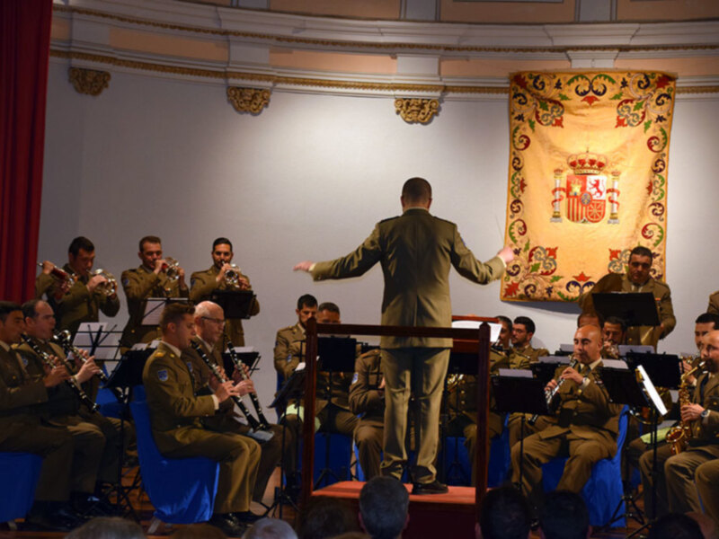 Orquesta militar en un escenario con un fondo decorado con un león y corona, bajo la dirección de un músico en el centro.