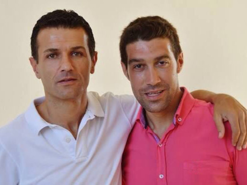 Dos hombres posando juntos en una foto profesional, uno con camisa blanca y el otro con camisa rosa.