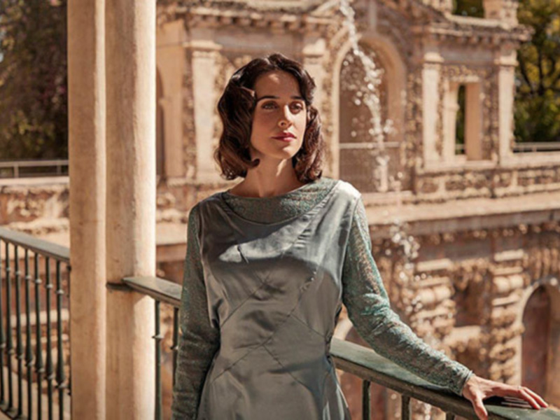 Mujer elegante en vestido de seda en un balcón con arquitectura histórica al fondo.