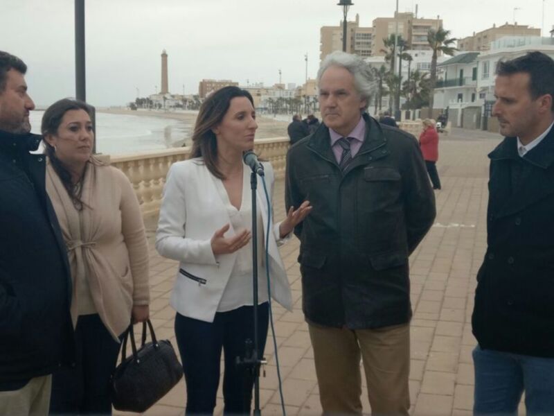 Una mujer habla en un micrófono mientras dos hombres la observan en una calle con edificios y un monumento en el fondo.