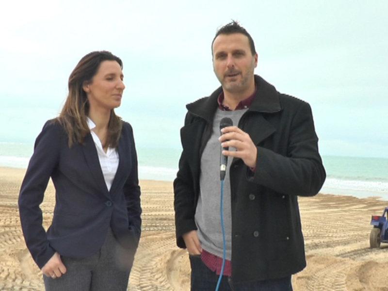 Entrevista en la playa con un reportero y una presentadora, con maquinaria de construcción al fondo.