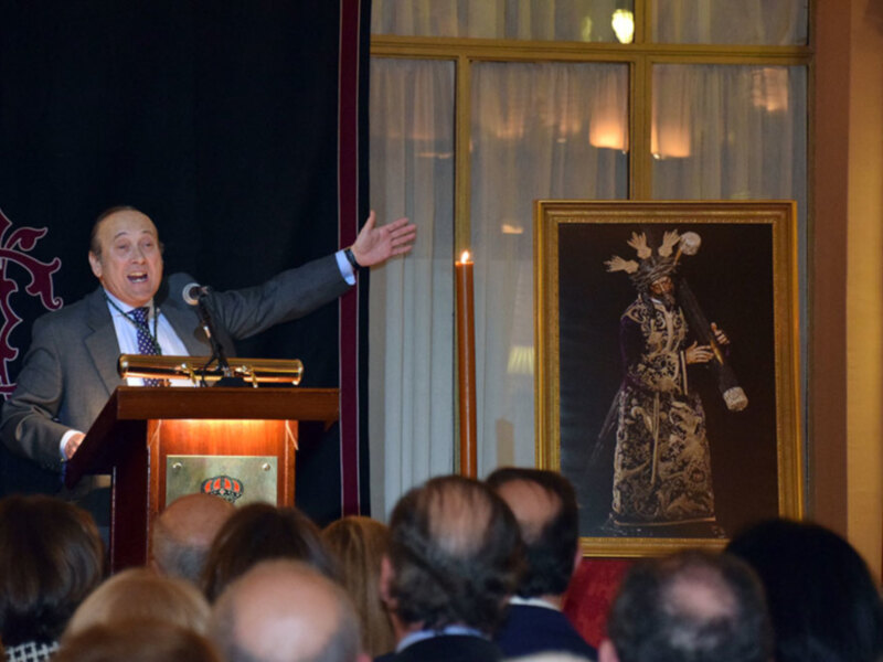 Un hombre habla en un evento formal, detrás de él hay una pintura de un personaje vestido con traje y corona.