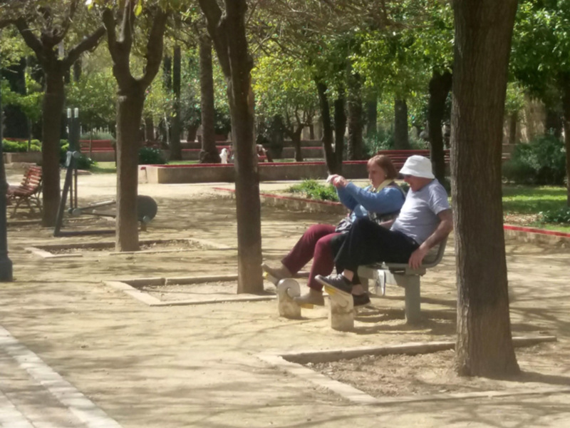 Dos personas descansando en un banco de un parque con árboles y bancos alrededor.