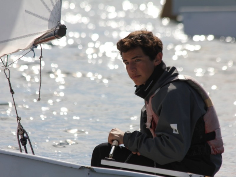 Un joven a bordo de un barco, con el viento soplando su cabello y la reflexión del agua en el fondo.